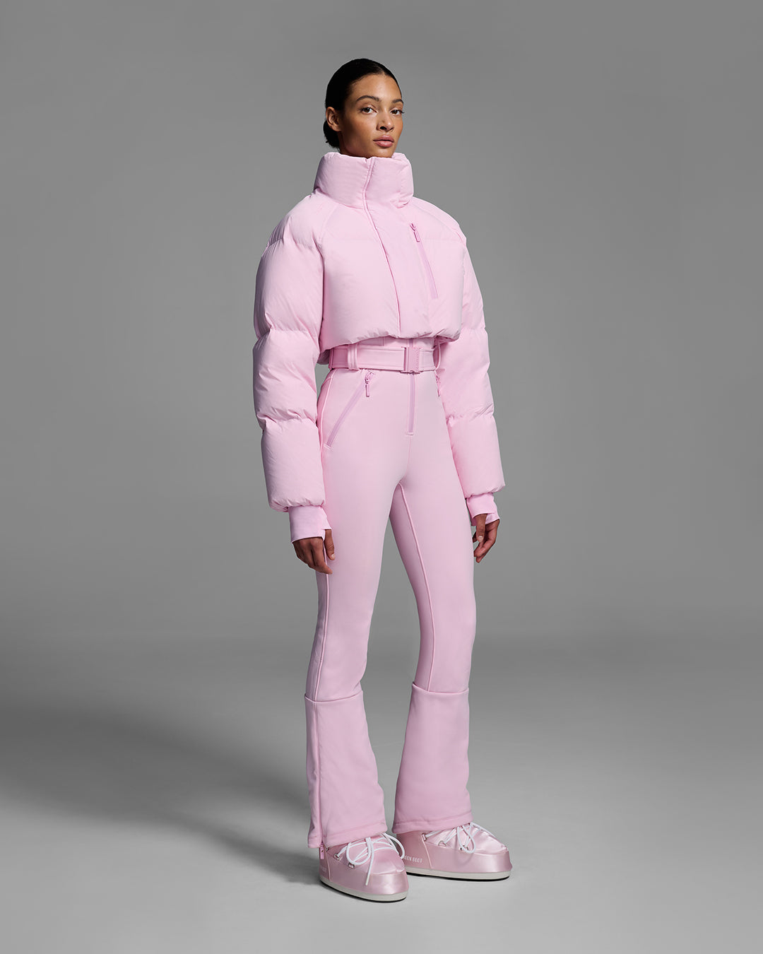 Female model wearing a peony pink ski suit with a puffer crop top, belted waist, and 4-way stretch pant. #color_peony