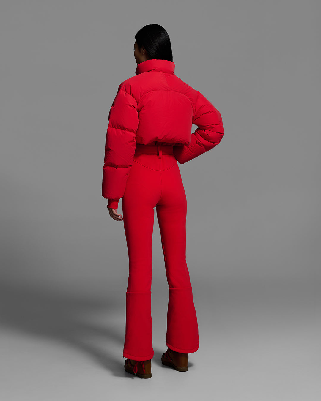 Back view of a female model wearing a bright poppy red ski suit with a puffer crop top, belted waist, and 4-way stretch pant. #color_poppy