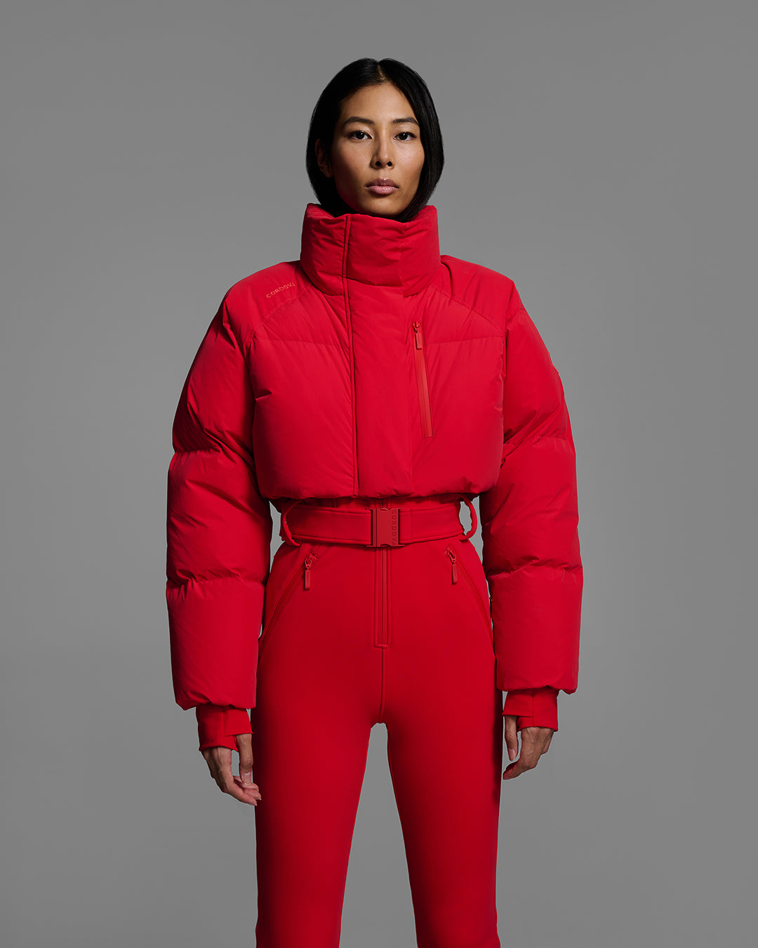 Detail front view of a female model wearing a bright poppy red ski suit with a puffer crop top, belted waist, and 4-way stretch pant. #color_poppy