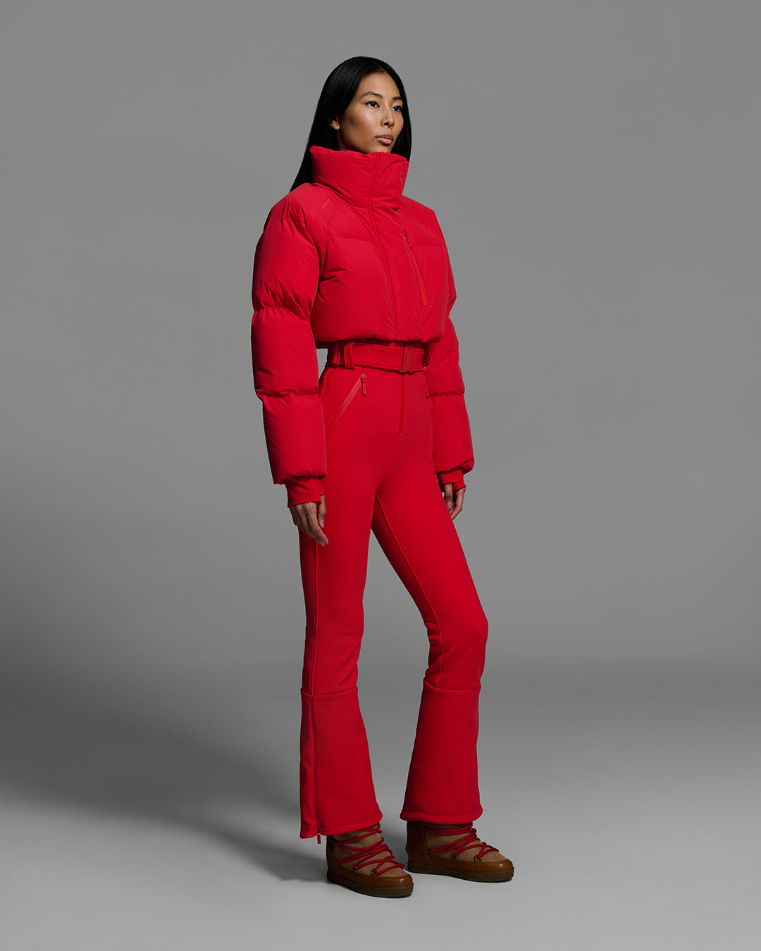 Female model wearing a bright poppy red ski suit with a puffer crop top, belted waist, and 4-way stretch pant. #color_poppy