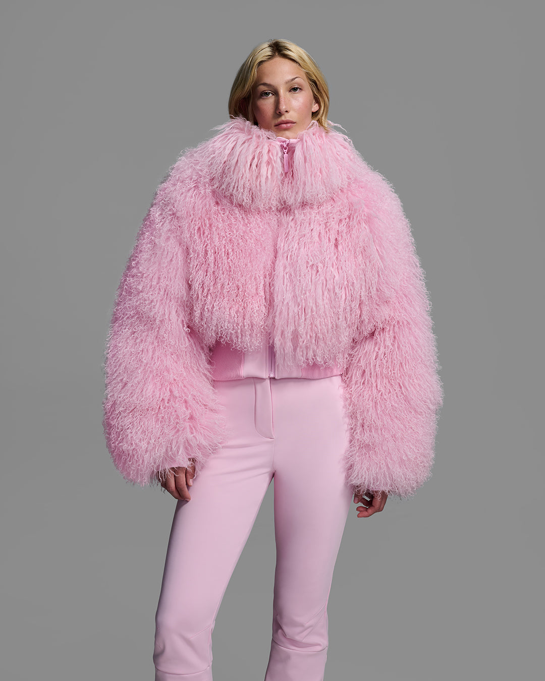 Female model wearing a natural shearling jacket in peony pink with matching ski pant. #color_peony