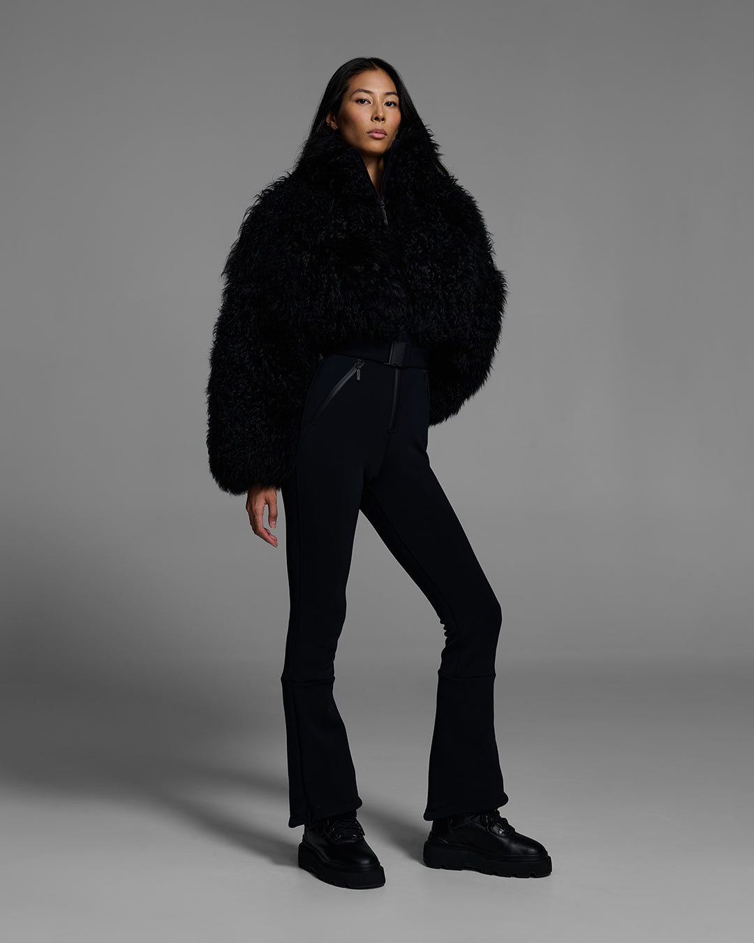 Woman wearing an onyx black color ski suit with a shearling top, belted waist, and 4-way stretch pant. #color_onyx