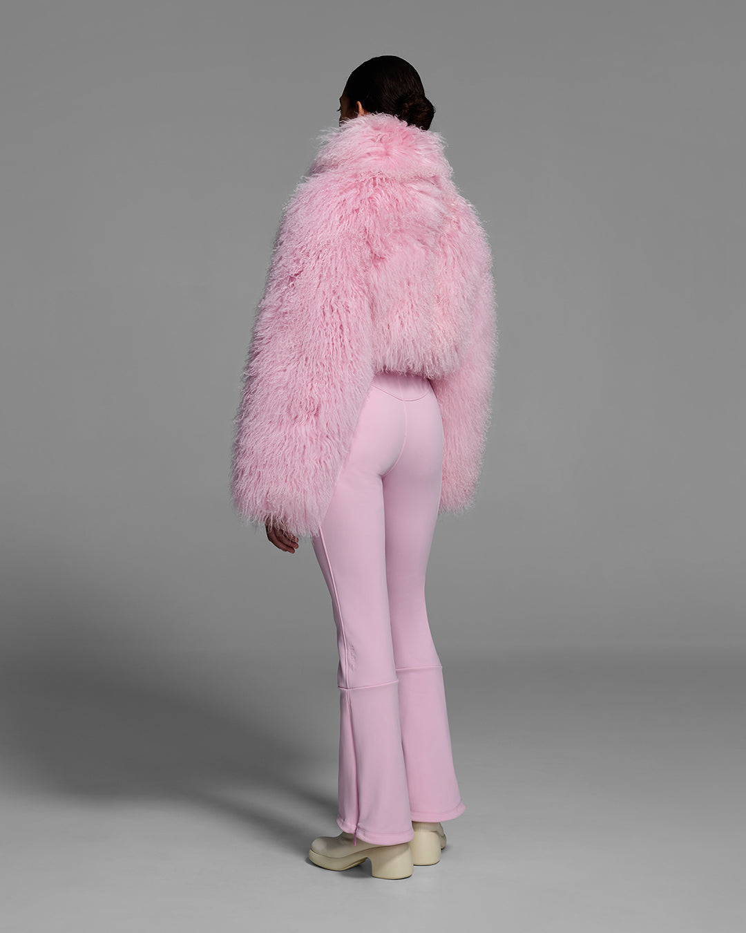 Back view of a woman wearing an peony pink color ski suit with a shearling top, belted waist, and 4-way stretch pant. #color_peony