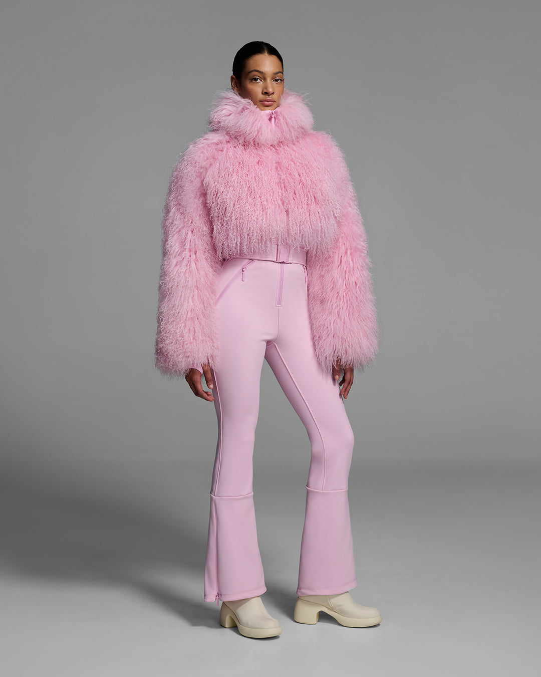 Woman wearing an peony pink color ski suit with a shearling top, belted waist, and 4-way stretch pant. #color_peony