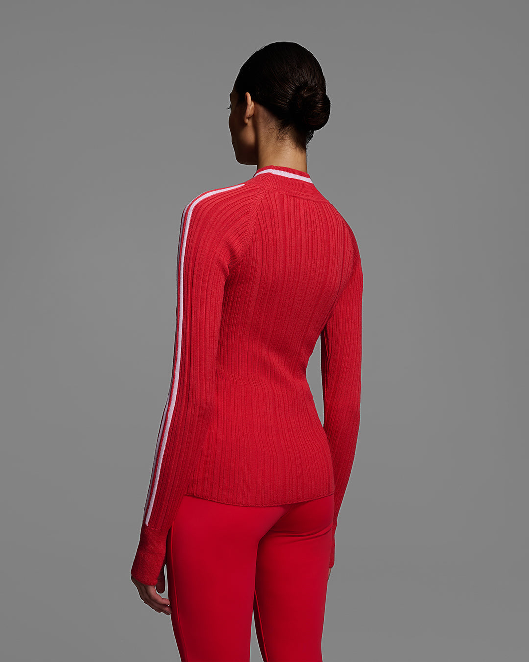 Back view of a woman wearing a bright poppy red ski sweater with racing stripes down the sleeves and on collar, with matching red ski pants. #color_poppy