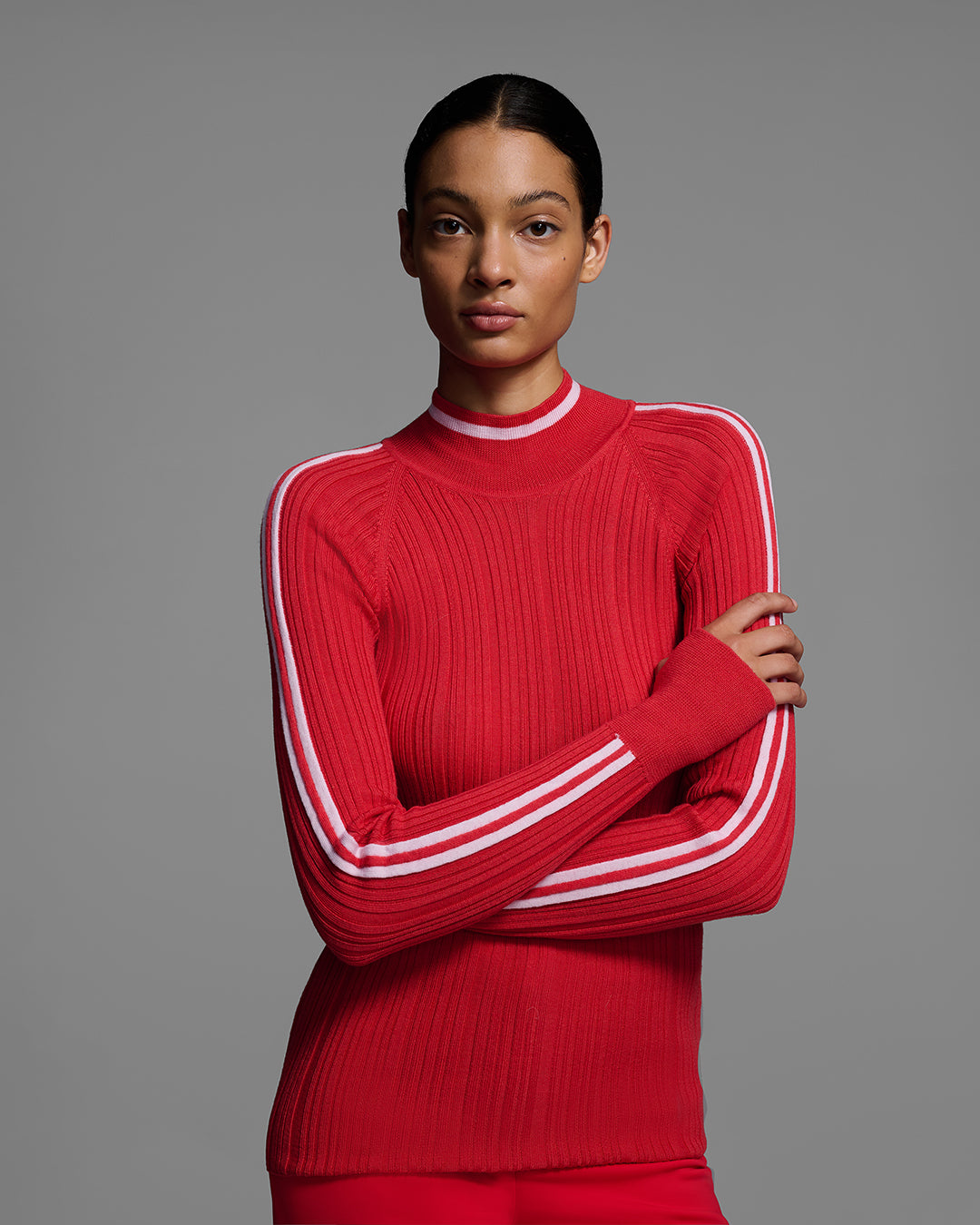 Woman wearing a bright poppy red ski sweater with racing stripes down the sleeves and on collar, with arms crossed. #color_poppy