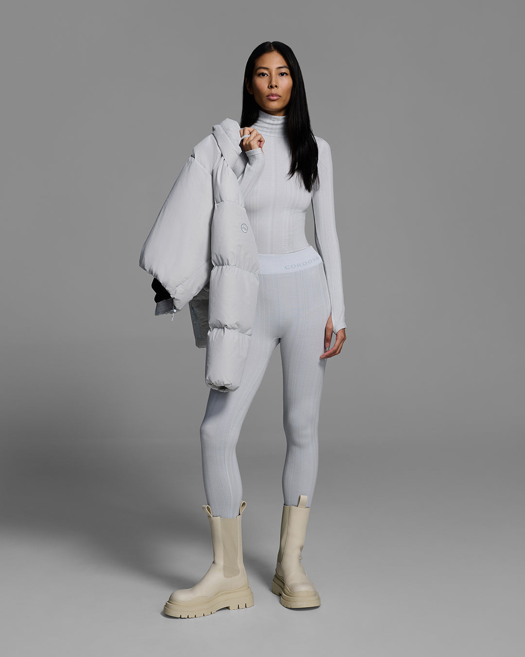 Woman wearing a smoke gray color turtleneck and matching pant base layer set made of merino wool blend, holding a matching puffer jacket over her shoulder. #color_smoke