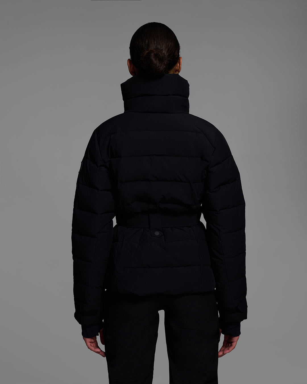 Back view of a woman wearing a belted down-fill puffer ski jacket in onyx black with matching pant and ski boots. #color_onyx