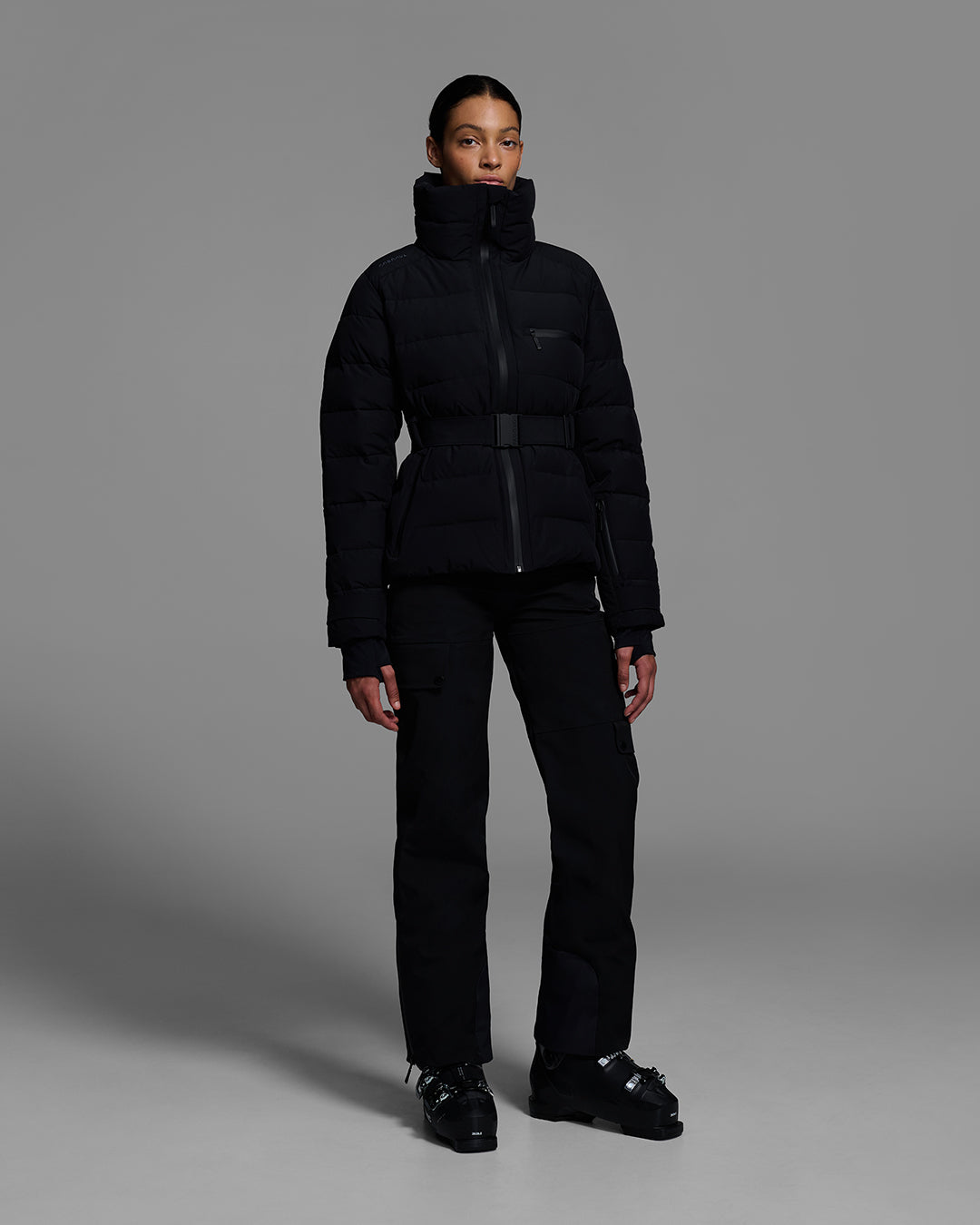 Woman wearing a belted down-fill puffer ski jacket in onyx black with matching pant and ski boots. #color_onyx