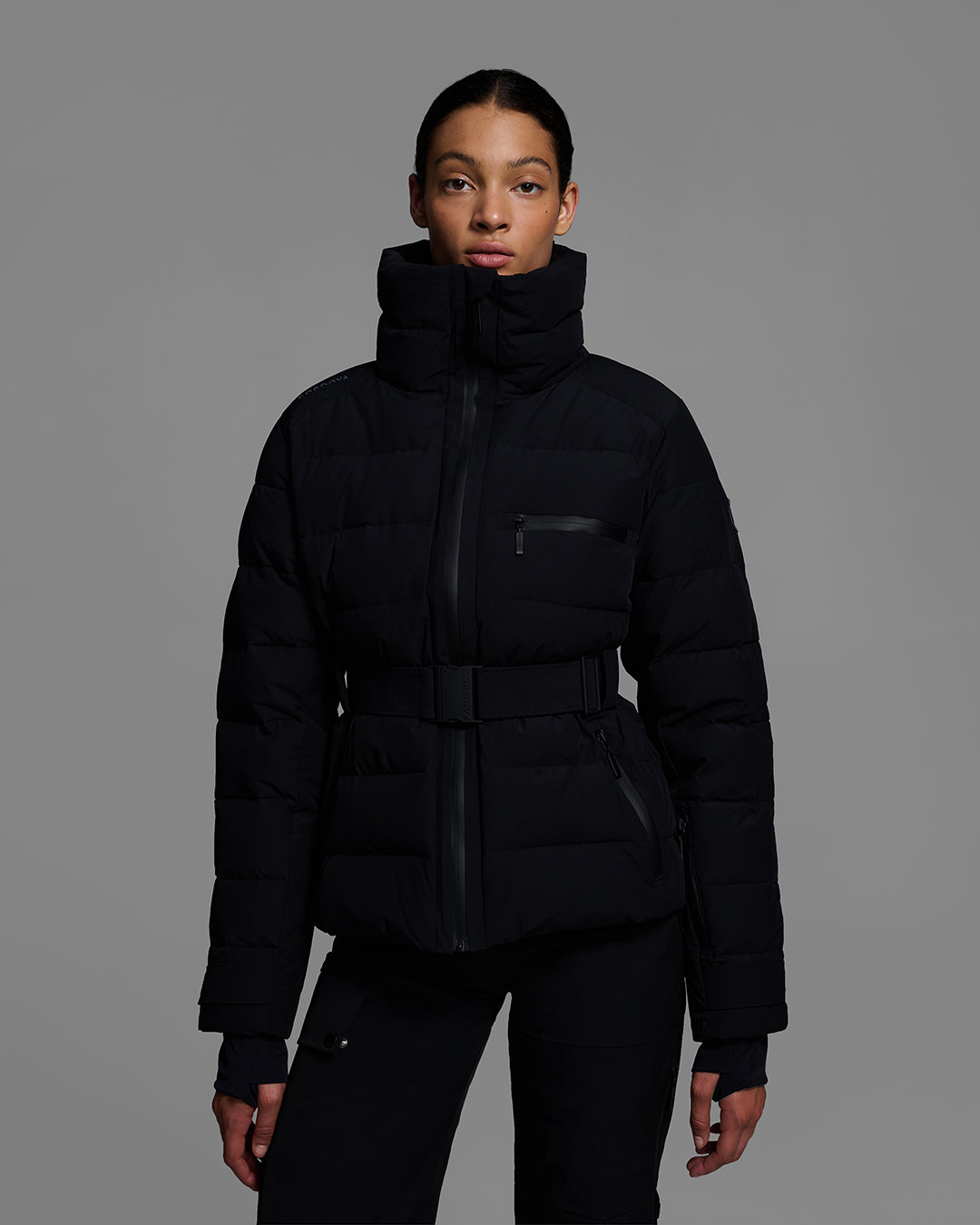 Front detail view of a woman wearing a belted down-fill puffer ski jacket in onyx black with matching pant and ski boots. #color_onyx