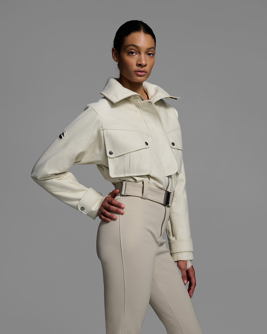 Detail front view of a woman wearing a natural white color wool top ski suit with belted waist and tan color 4-way stretch bottom. #color_natural / sand