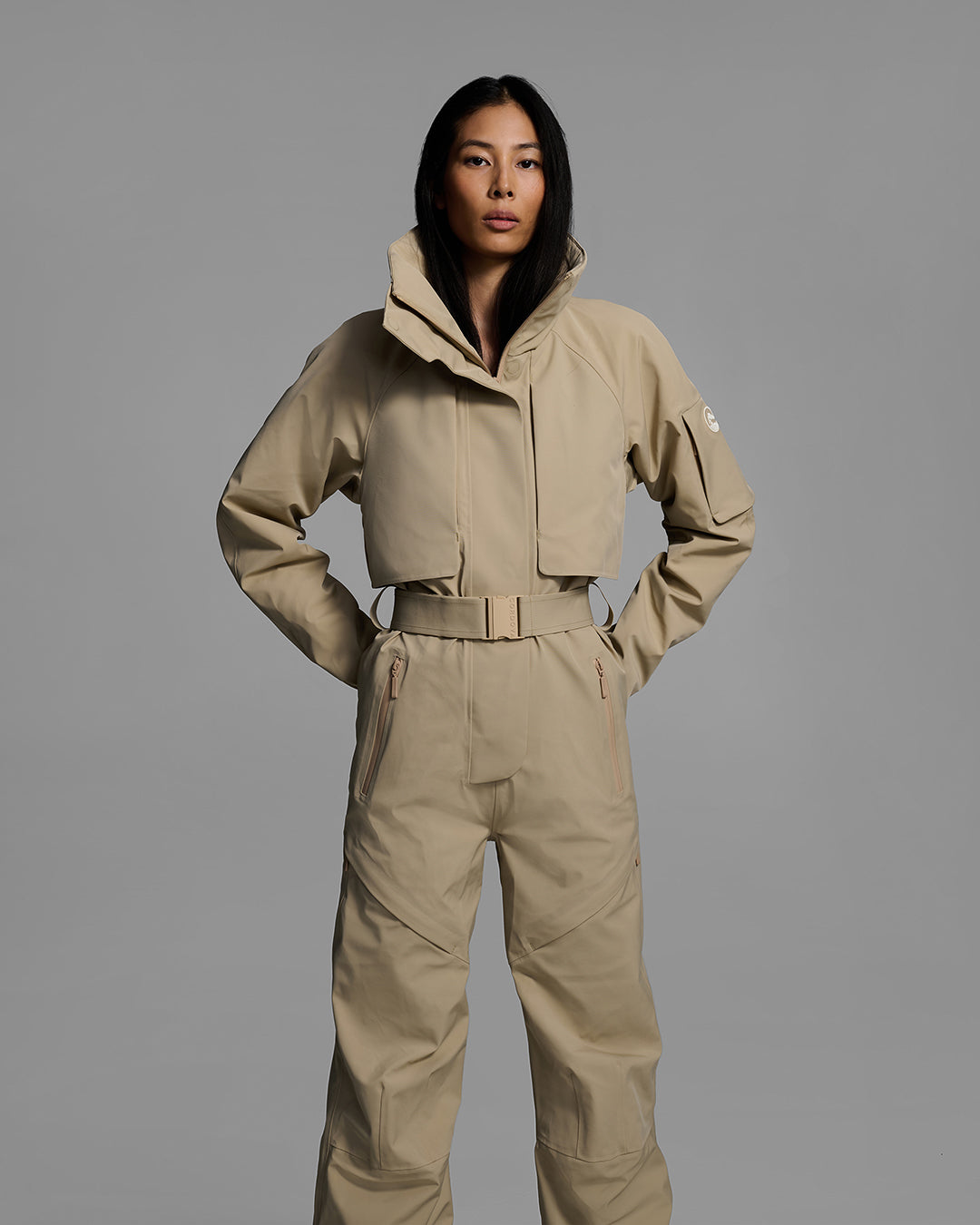Front view of a woman wearing a tan color waterproof technical fabric ski suit with a belted waist. #color_sand