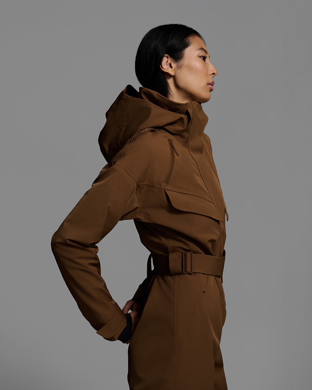 Side detail view of a female model wearing a belted sepia brown color ski suit made of technical waterproof fabric and zippers. #color_sepia
