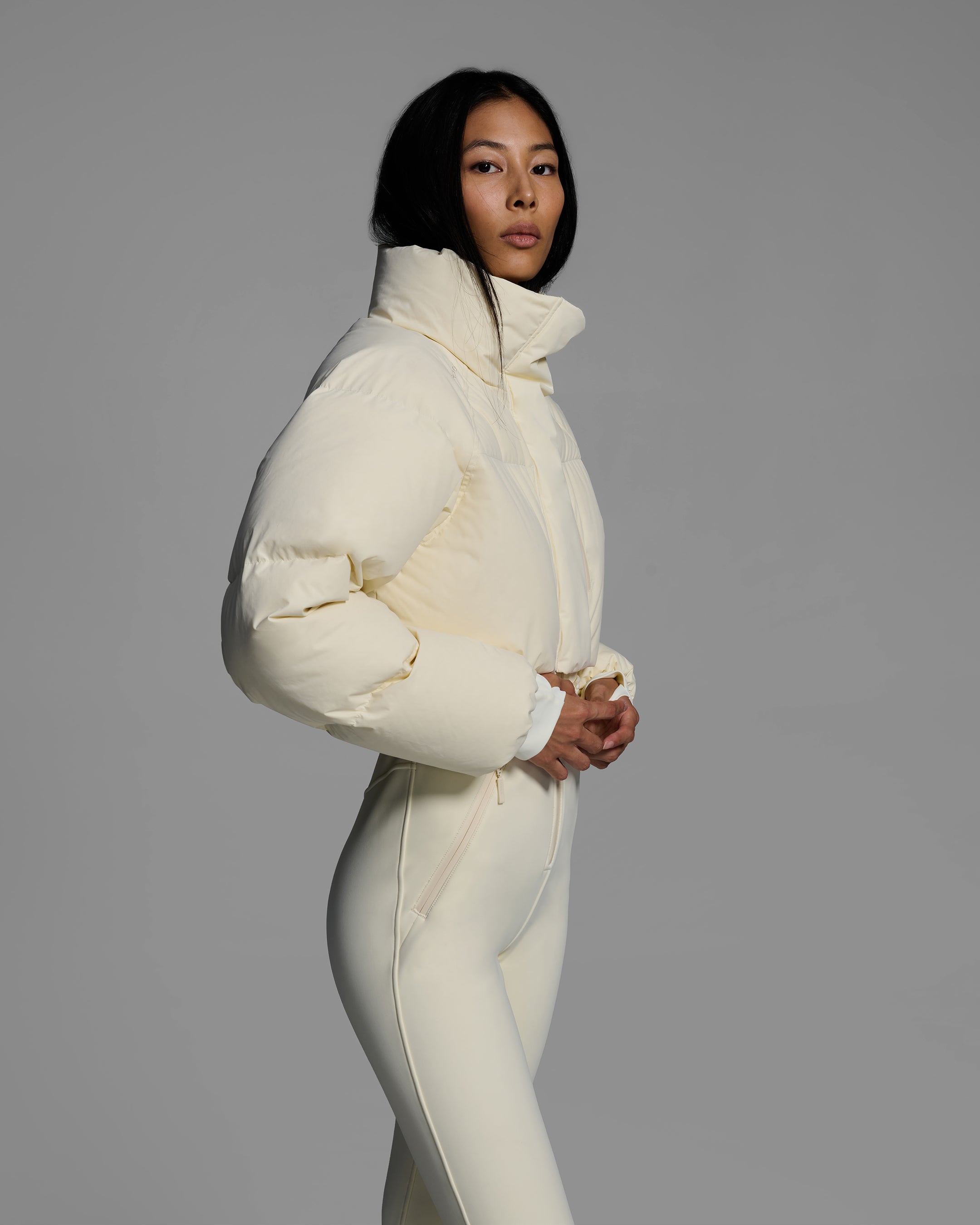Female model wearing an alabaster white ski suit with a white puffer crop top, belted waist, and 4-way stretch pant. #color_alabaster