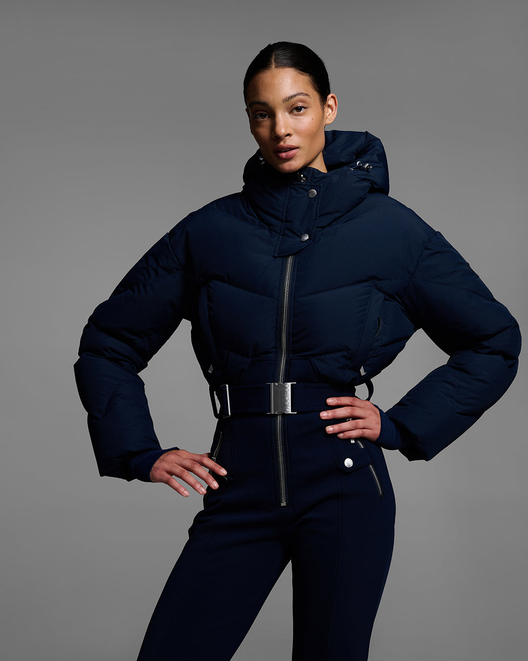 Female model with hands on hips wearing a belted indigo blue color ski suit with down filled top and 4-way stretch bottom. #color_indigo