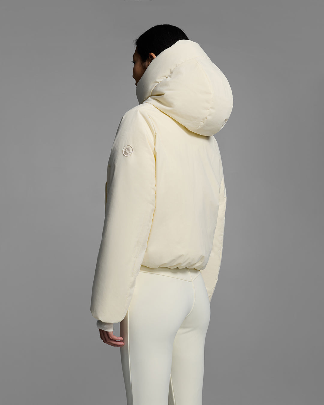 Back view of a female model wearing a down filled ski jacket in alabaster off-white fabric with a matching off-white ski pant. #color_alabaster
