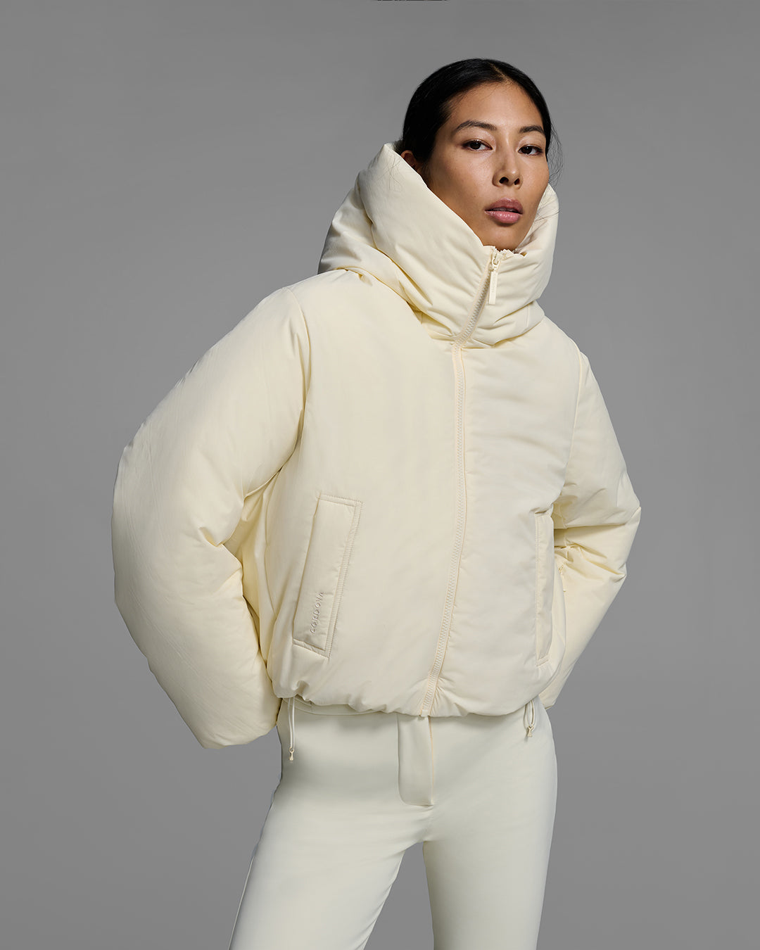 Detail view of a female model with her hands on her hips wearing a down filled ski jacket in alabaster off-white fabric, zipped up with a matching off-white ski pant. #color_alabaster