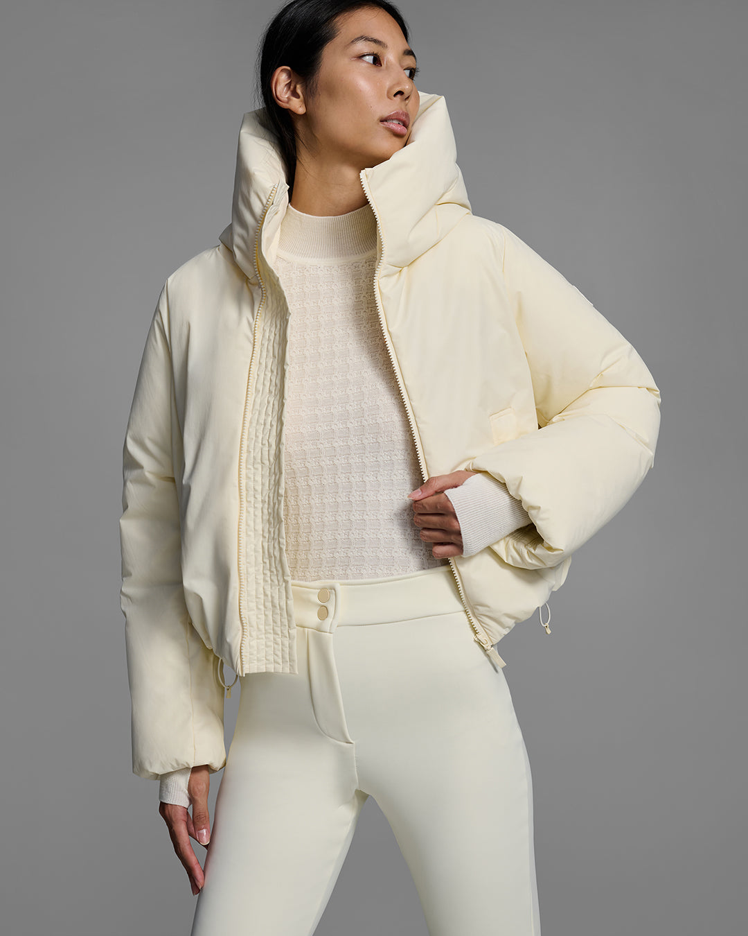 Detail view of a female model wearing a down filled ski jacket in alabaster off-white fabric, unzipped up with a matching off-white knit sweater and ski pant. #color_alabaster
