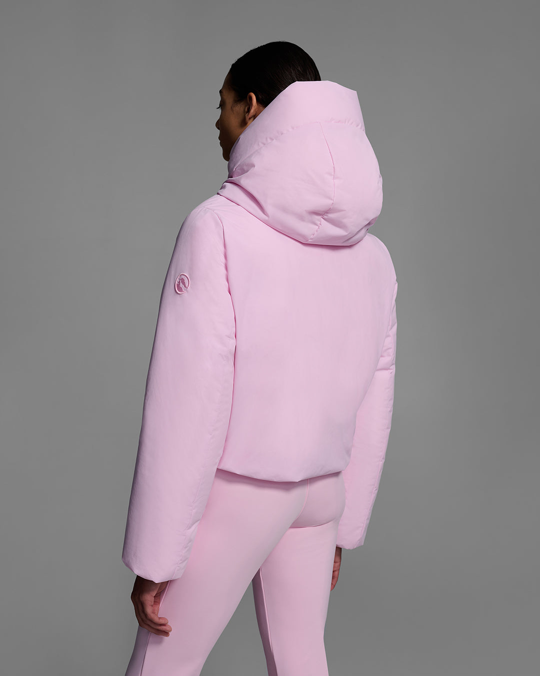 Back view of a female model wearing a down filled ski jacket in peony pink fabric with a matching pink ski pant. #color_peony