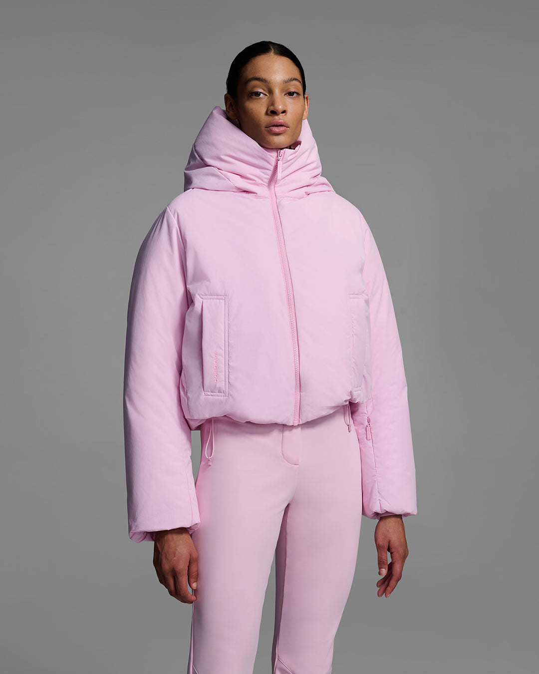 A detail view of a female model wearing a down filled ski jacket in peony pink fabric, zipped up with a matching pink ski pant. #color_peony