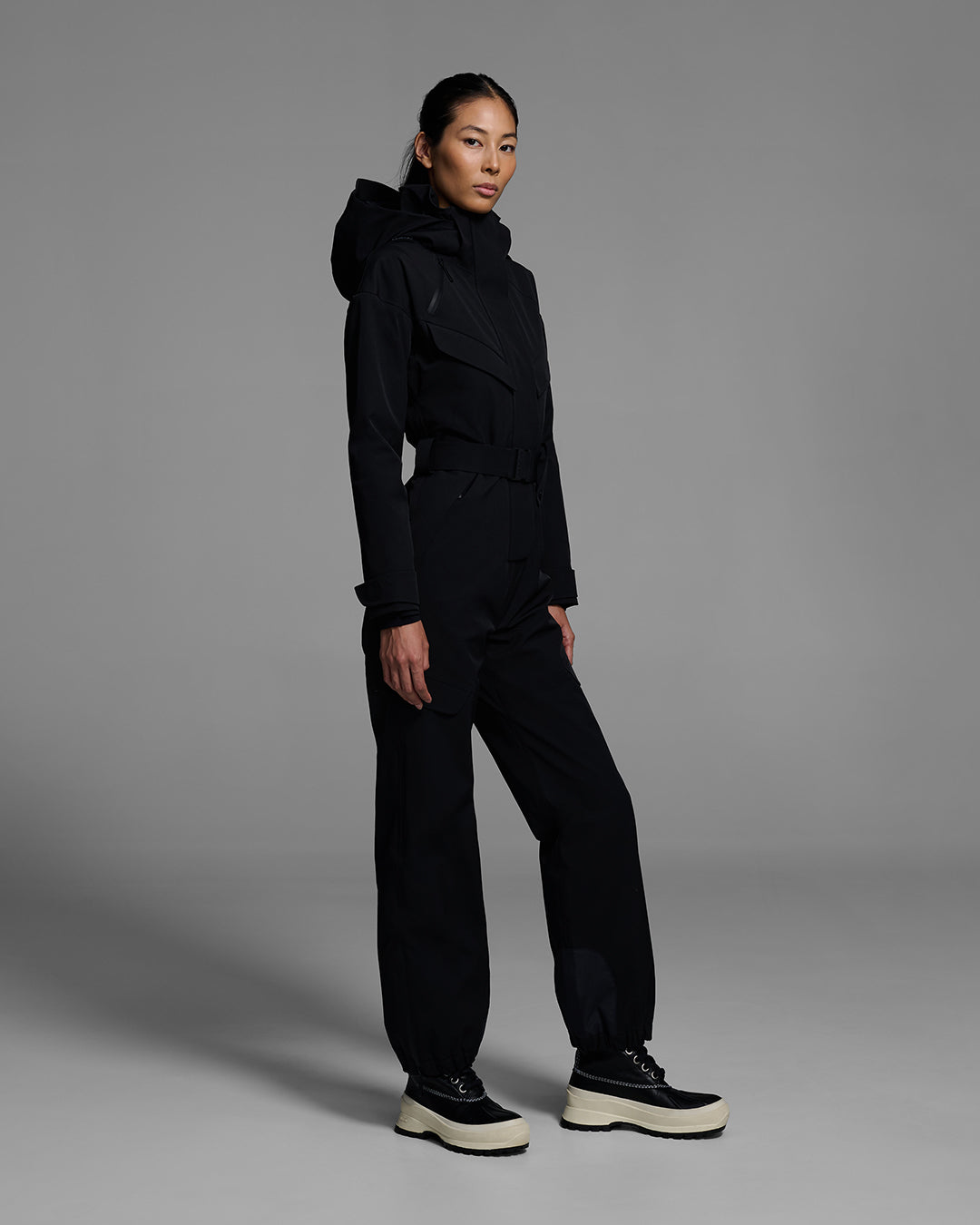 Female model wearing a belted onyx black color ski suit made of technical waterproof fabric with  zippers. #color_onyx