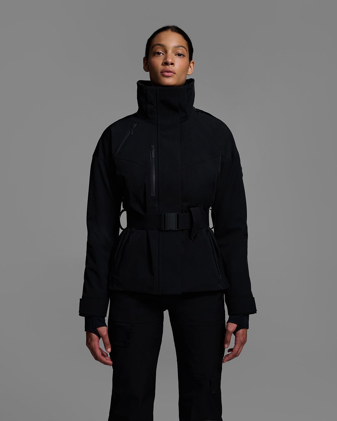 Female model wearing a belted ski jacket in onyx black made of technical waterproof fabric with waterproof zippers, and a matching black pant. #color_onyx
