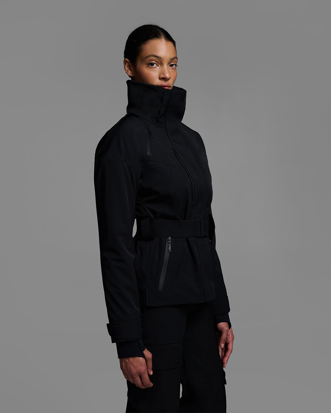 Side view of a female model wearing a belted ski jacket in onyx black made of technical waterproof fabric with waterproof zippers, and a matching black pant. #color_onyx