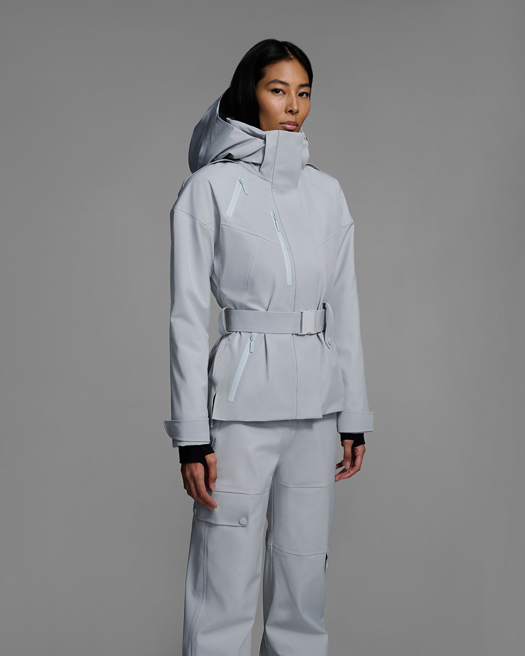 Detail shot of a female model wearing a belted ski jacket in smoke gray technical waterproof fabric with waterproof zippers, and a matching gray pant. #color_smoke