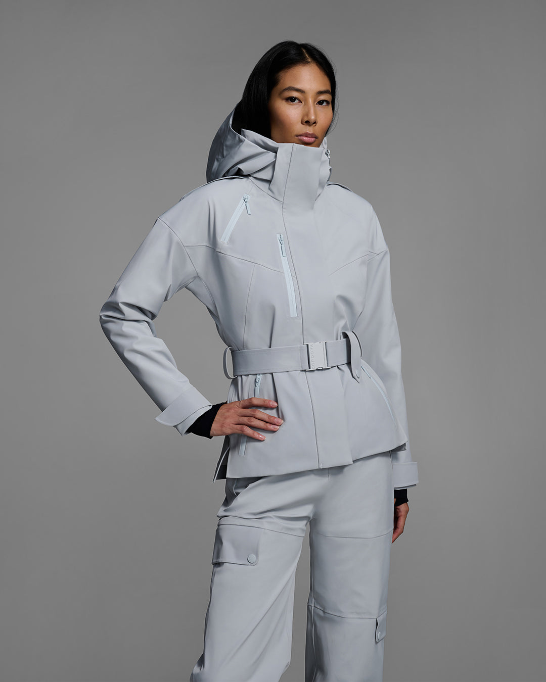Female model wearing a belted ski jacket in smoke gray technical waterproof fabric with waterproof zippers, and a matching gray pant. #color_smoke