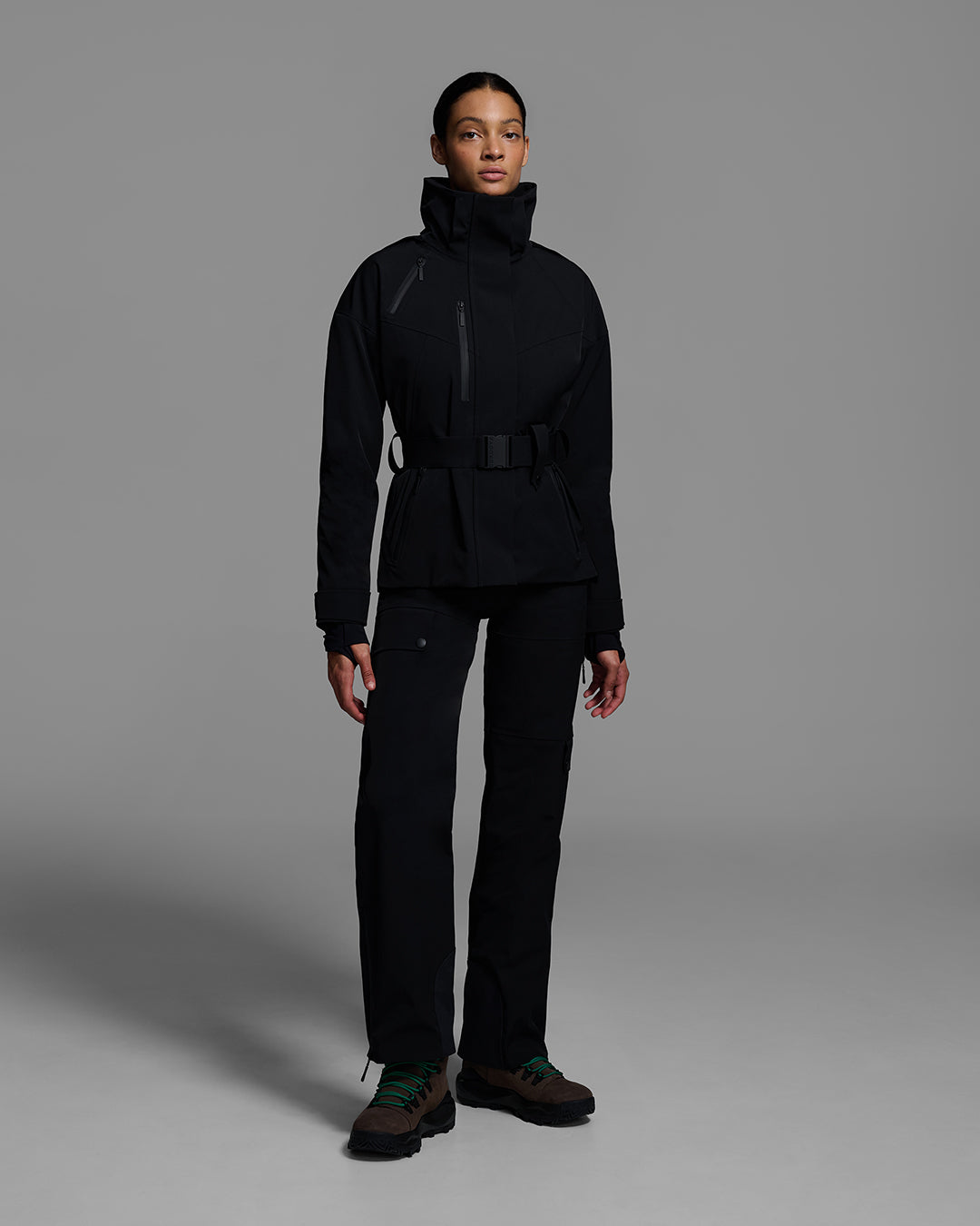 Female model wearing a belted ski jacket in onyx black made of technical waterproof fabric with waterproof zippers, and a matching black pant. #color_onyx