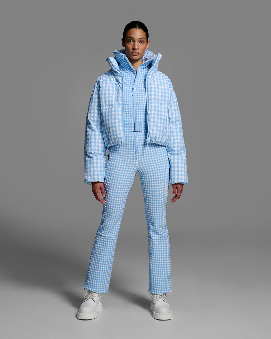 Female model wearing a belted cerulean blue and white gingham print ski suit with matching down jacket, unzipped. #color_cerulean gingham
