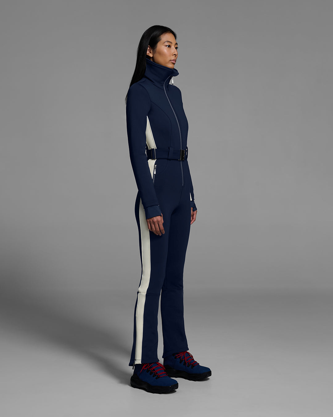 Female model wearing a body hugging ski suit in indigo blue with a white side stripe and belted waist. #color_indigo