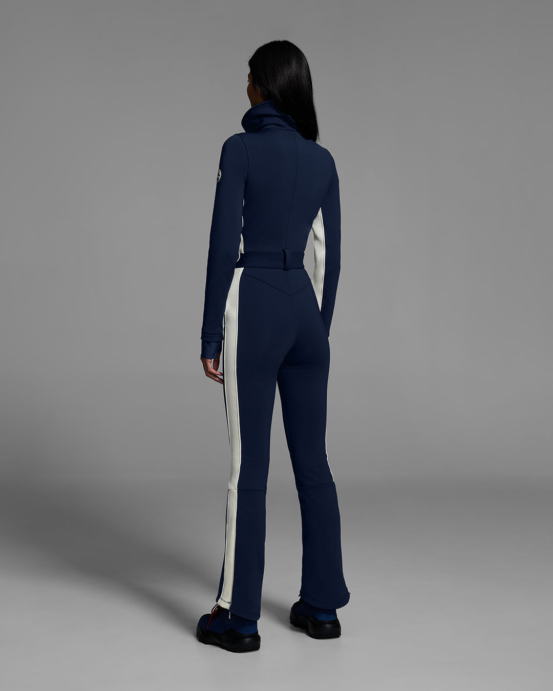 Back view of a female model wearing a body hugging ski suit in indigo blue with a white side stripe and belted waist. #color_indigo
