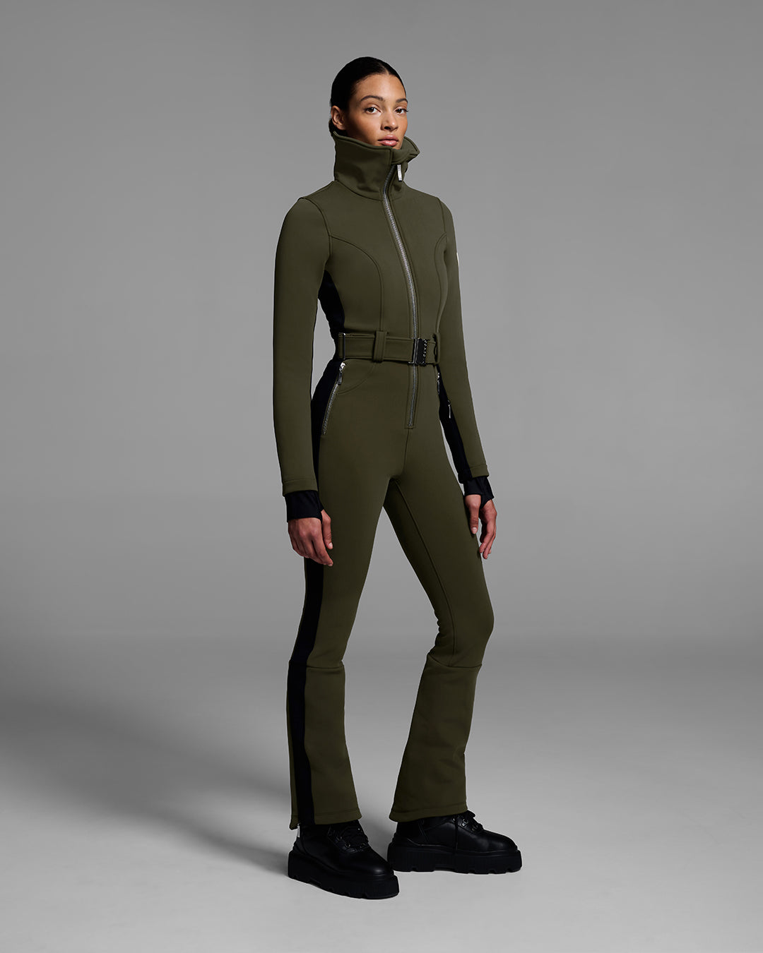 Female model wearing a body hugging ski suit in khaki green with a black side stripe and belted waist. #color_khaki