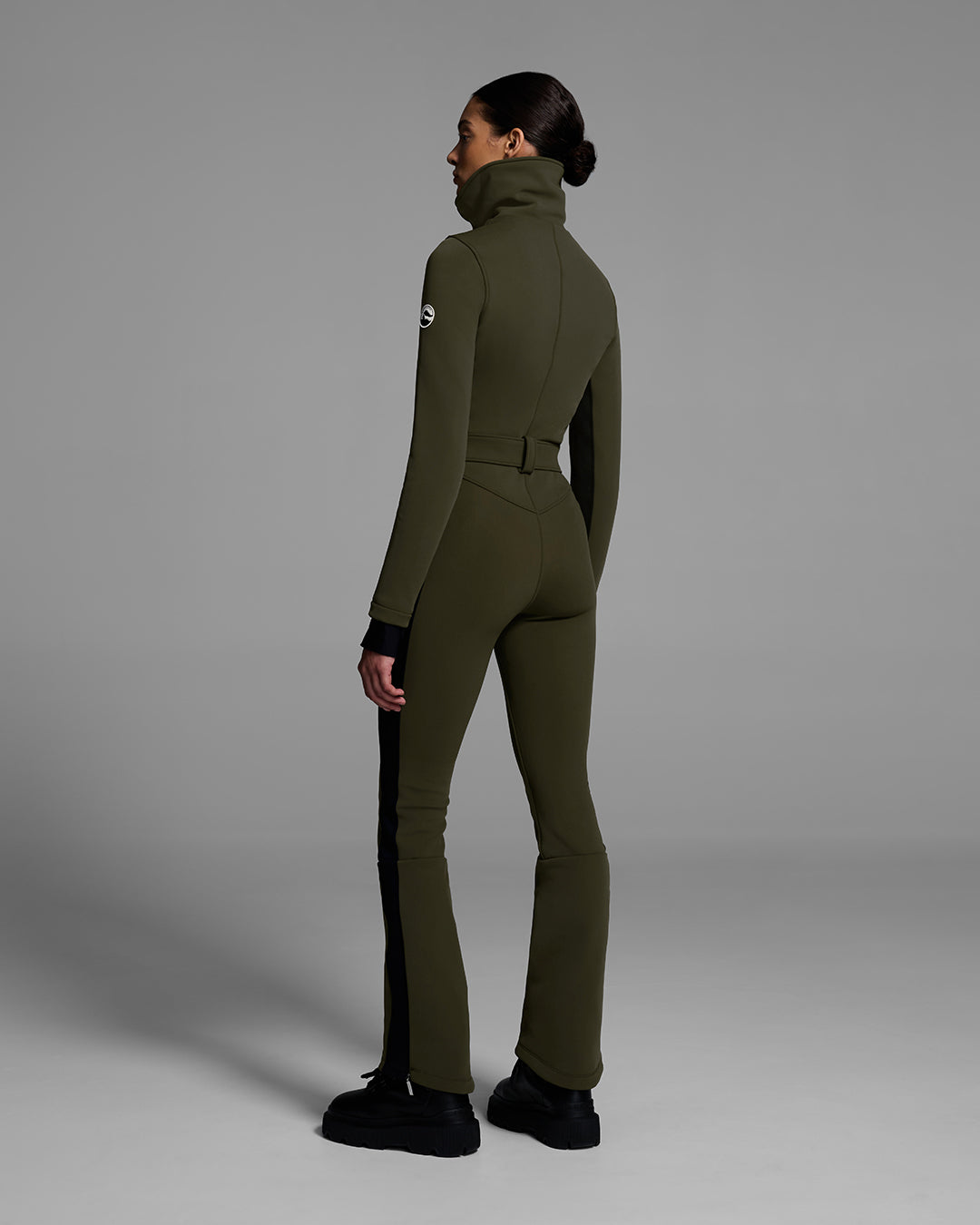 Back view of a female model wearing a body hugging ski suit in khaki green with a black side stripe and belted waist. #color_khaki