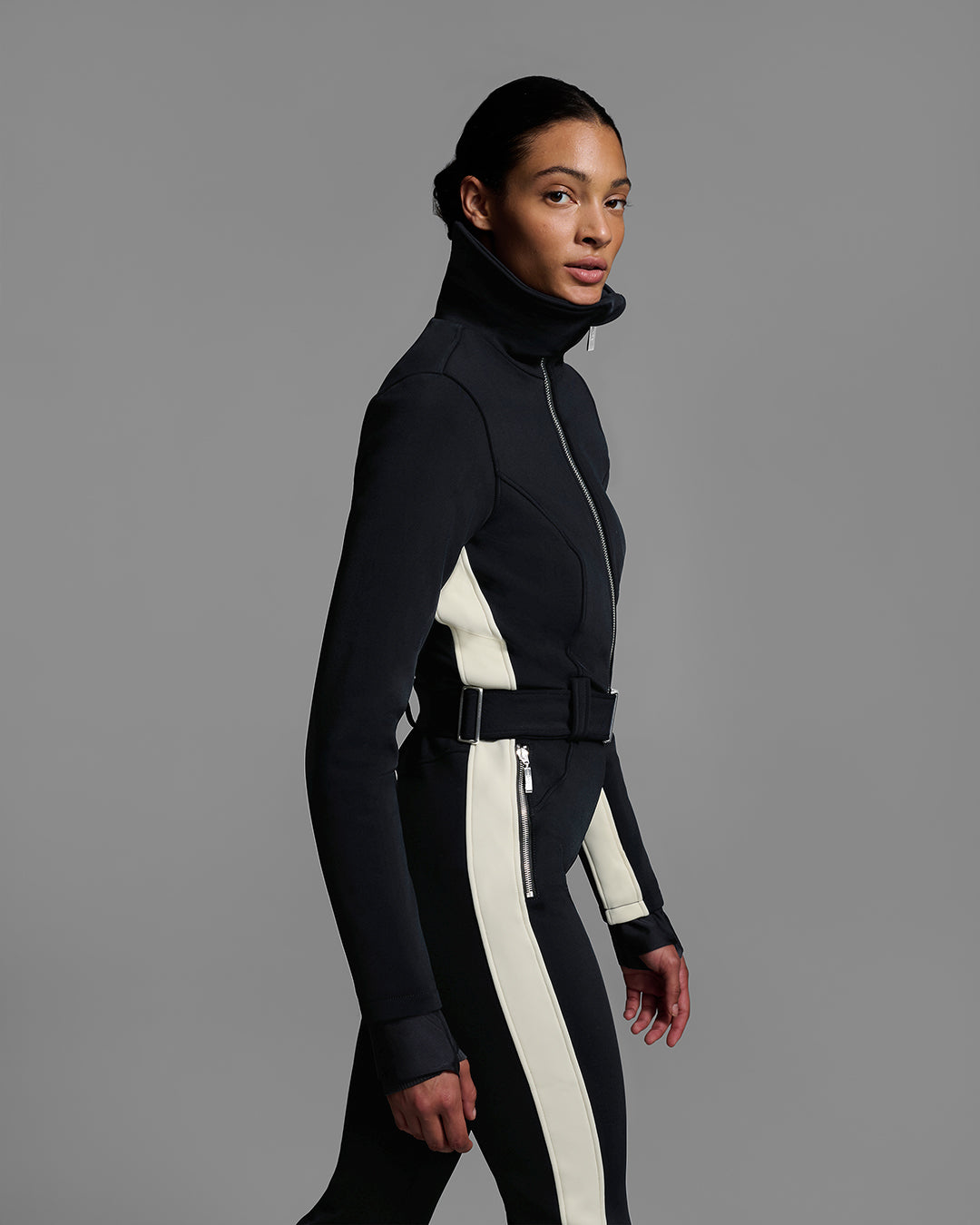 Detail side view of a female model wearing a body hugging ski suit in black with a white side stripe and belted waist. #color_onyx