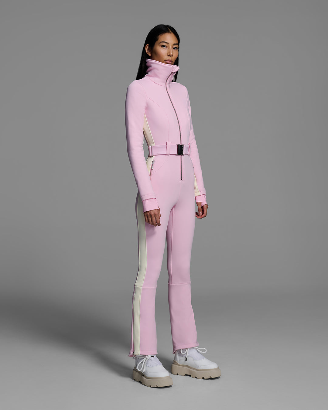 Female model wearing a body hugging ski suit in peony pink with a white side stripe and belted waist. #color_peony