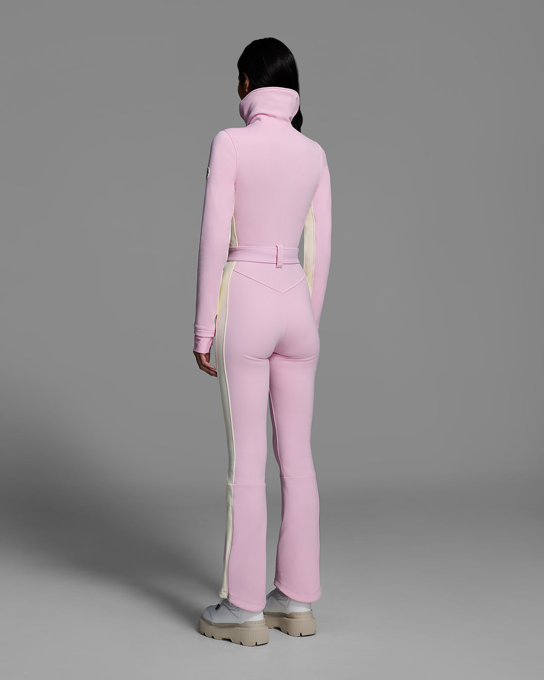 Back view of a a female model wearing a body hugging ski suit in peony pink with a white side stripe and belted waist. #color_peony