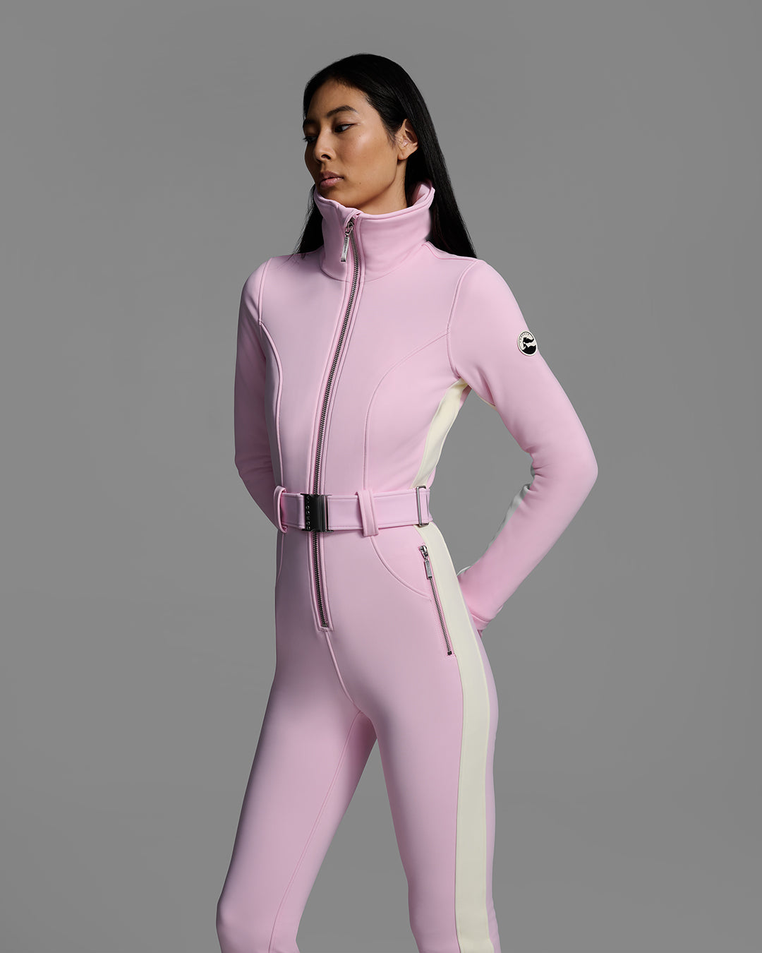 Detail view of a female model wearing a body hugging ski suit in peony pink with a white side stripe and belted waist. #color_peony