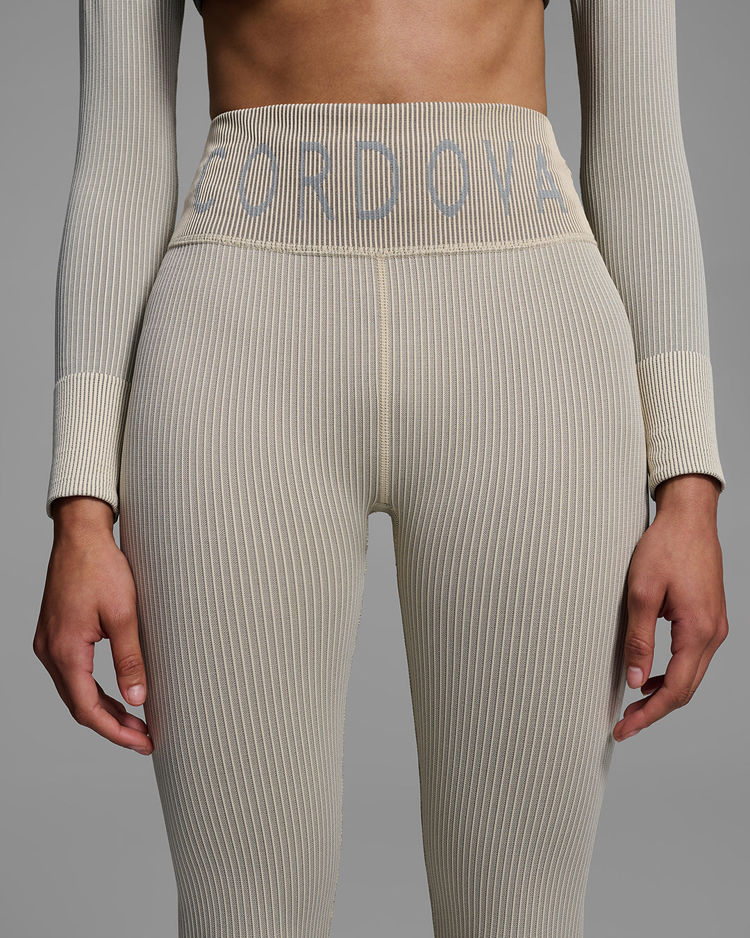 Detail view of a female model wearing a ski base layer pant in a compression knit fabric and ecru tan color with CORDOVA branding on the waist. #color_ecru