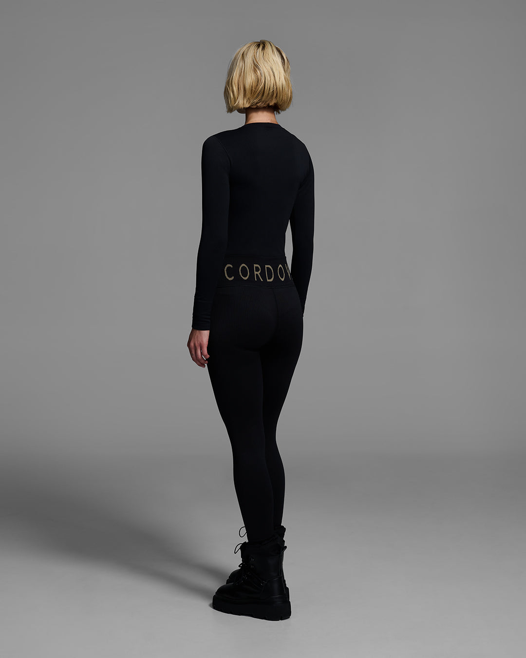 Back view of a female model wearing a matching ski base layer set in a compression knit fabric and onyx black color. #color_onyx