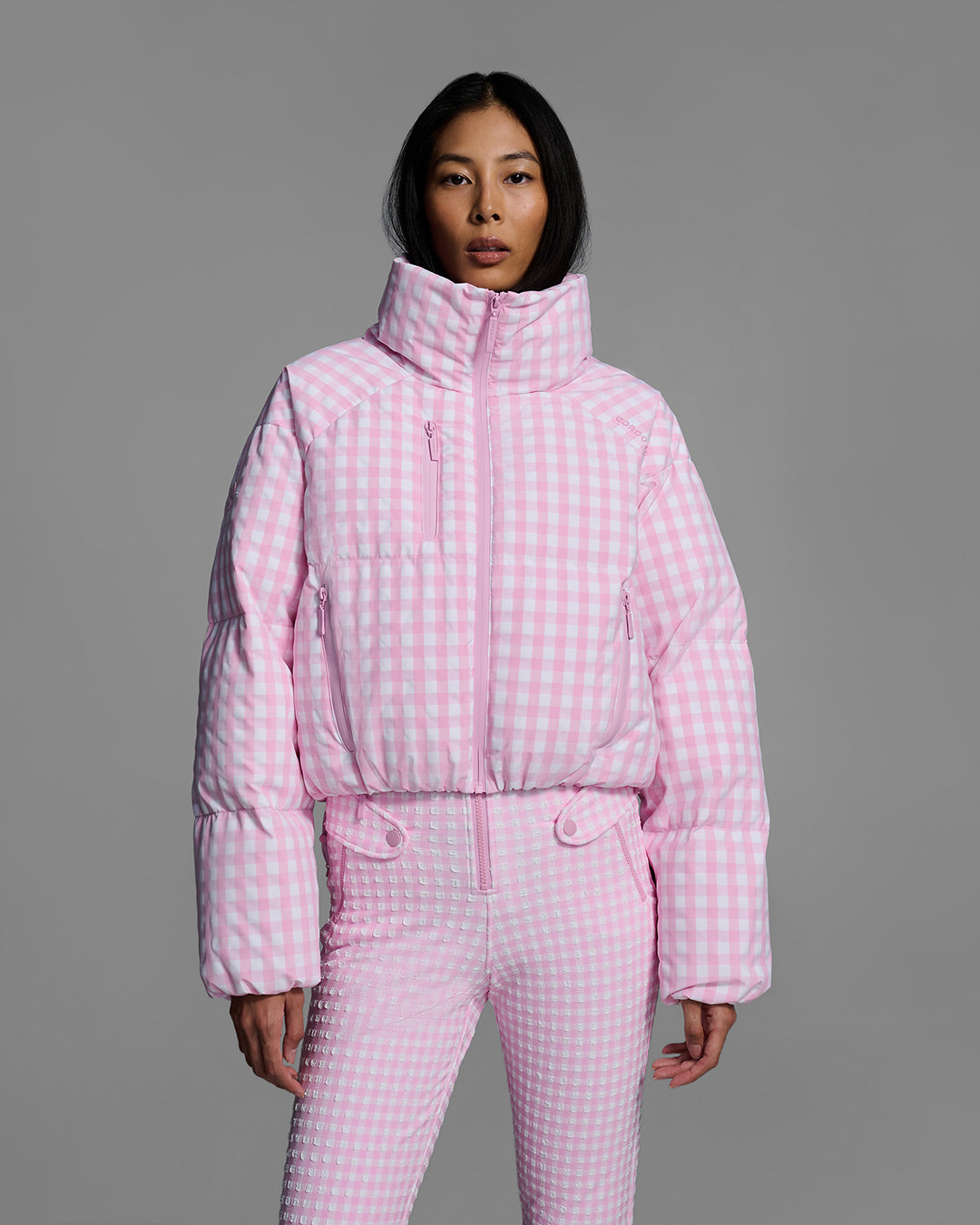 Detail view of a female model wearing a down filled ski jacket in peony pink gingham fabric with matching ski suit underneath. #color_peony gingham