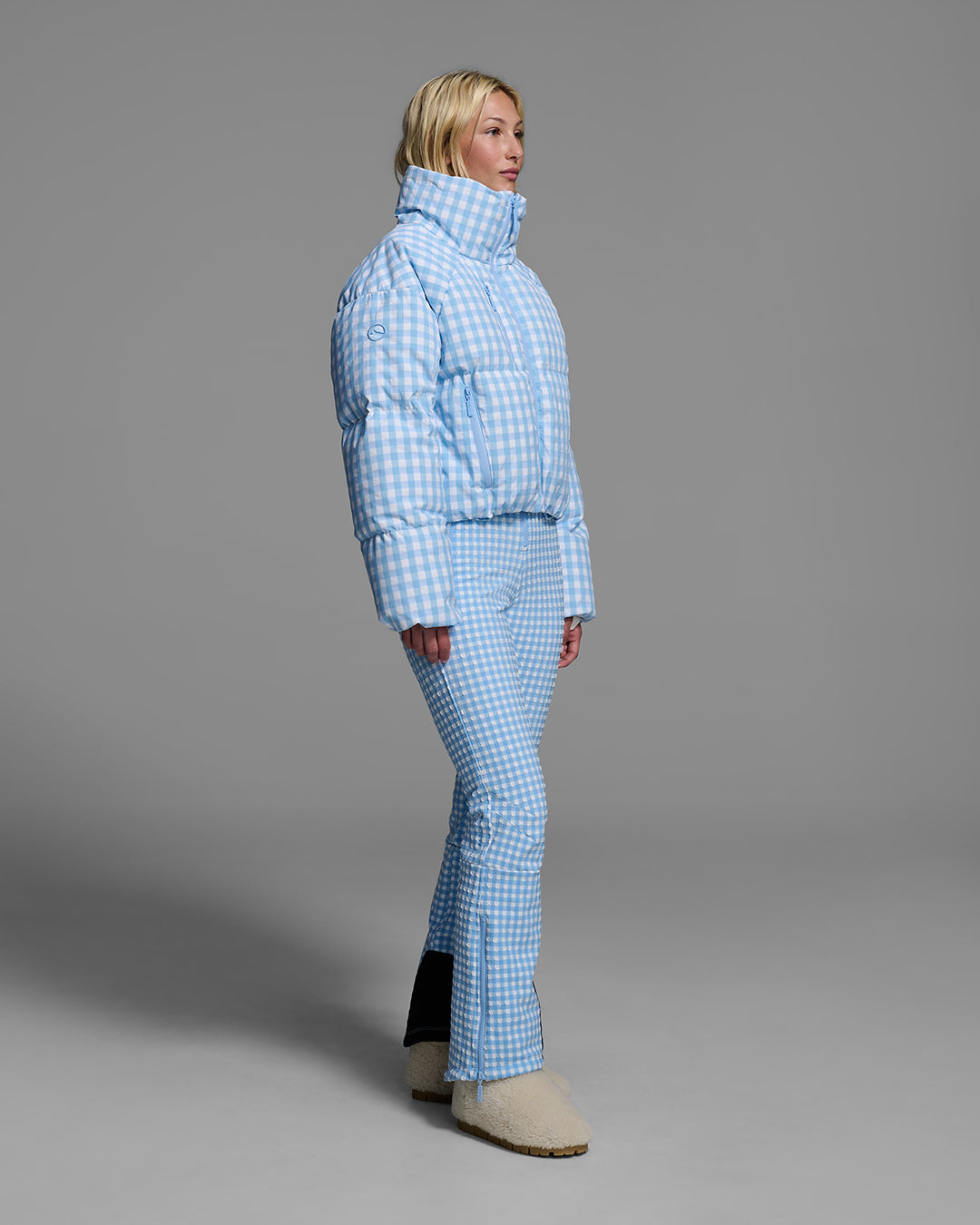 Female model wearing a down filled ski jacket in cerulean blue gingham fabric with matching ski suit underneath. #color_cerulean gingham