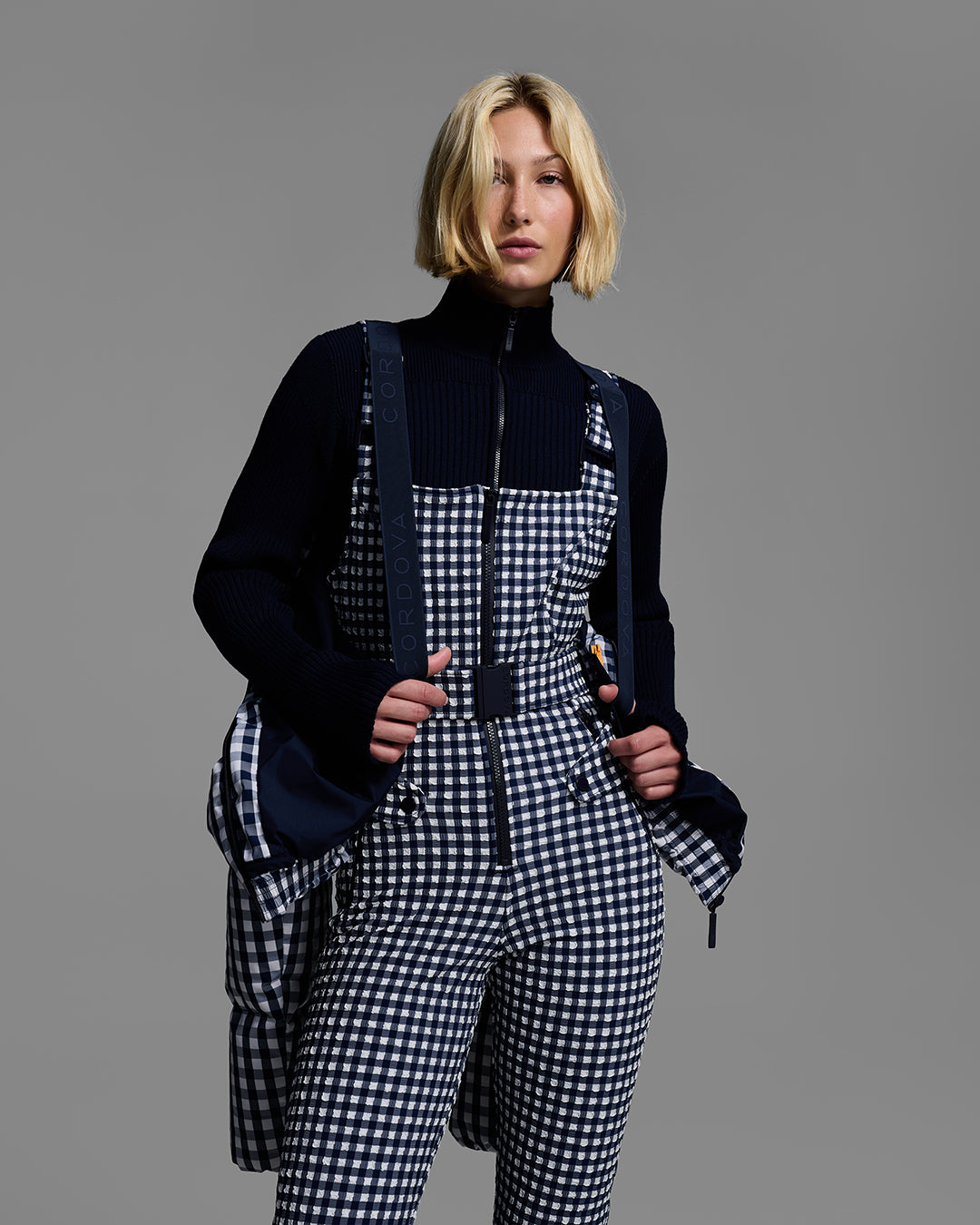 Female model wearing a down filled ski jacket in indigo blue gingham hanging off shoulders to highlight the backpack strap feature with a matching ski bib underneath. #color_indigo gingham