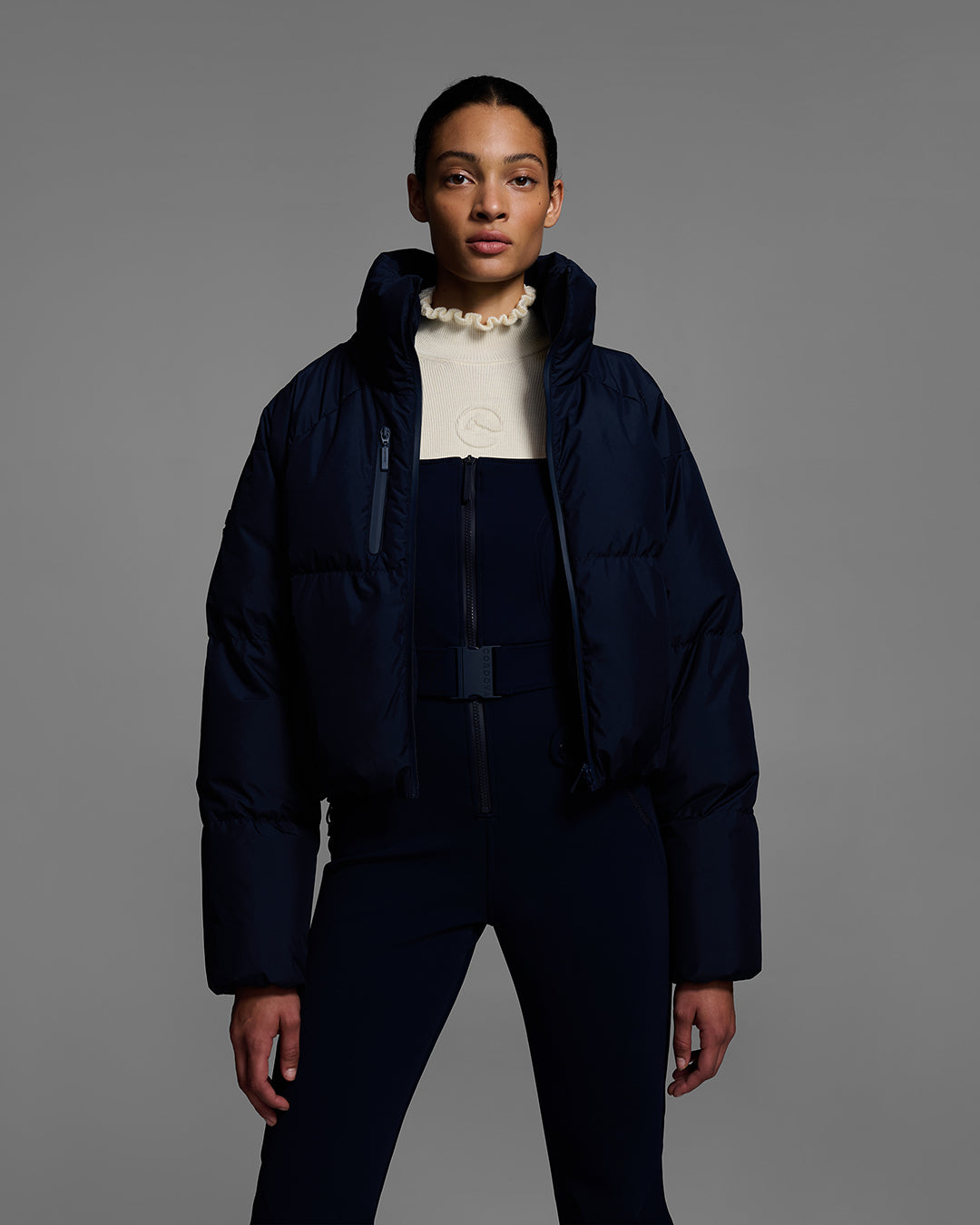 Female model wearing a down filled ski jacket in indigo blue fabric unzipped to show the matching ski bib beneath. #color_indigo