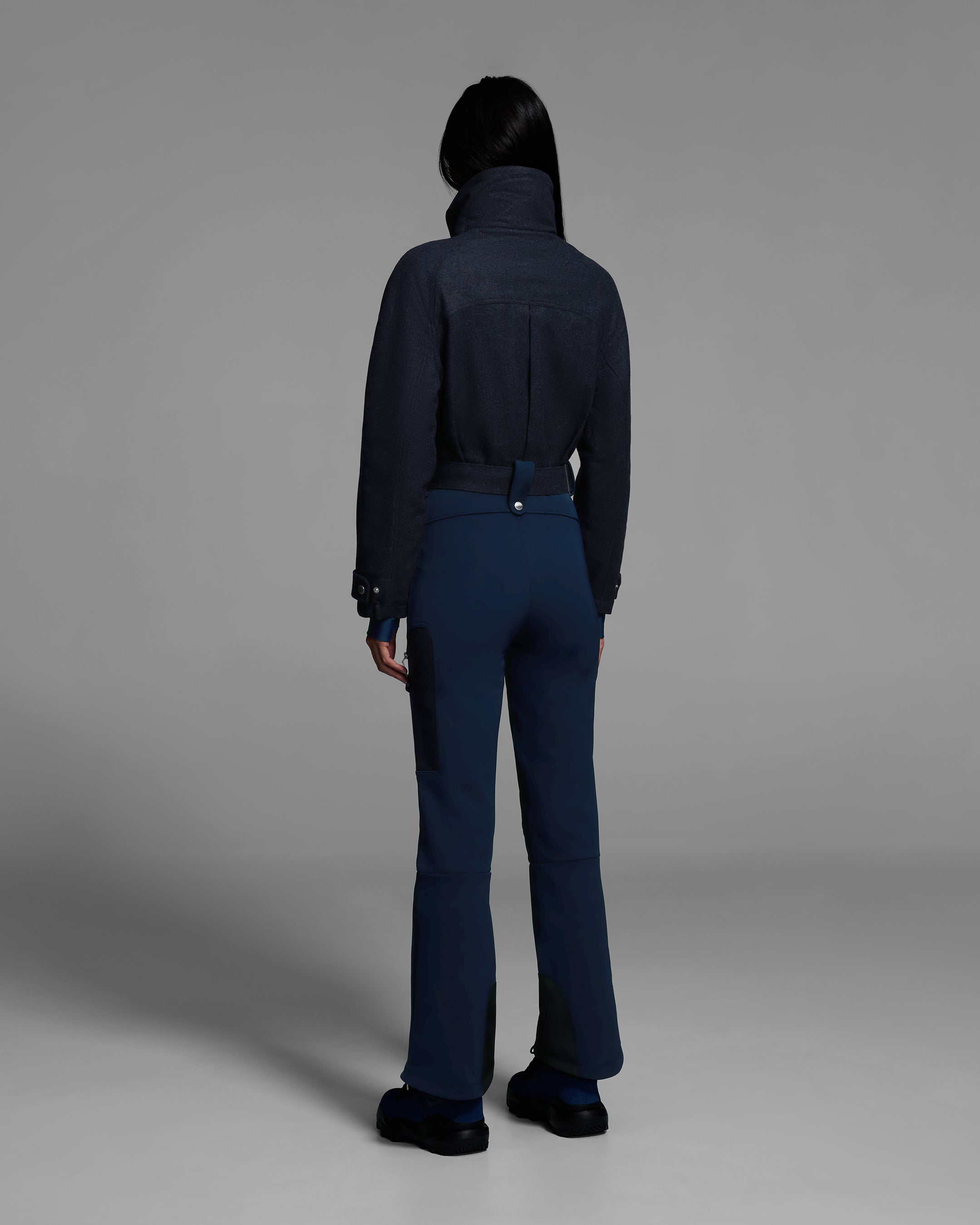 Back view of a female model wearing a belted indigo denim melange ski suit with a wool top and 4-way stretch water-resistant fabric bottom. #color_indigo