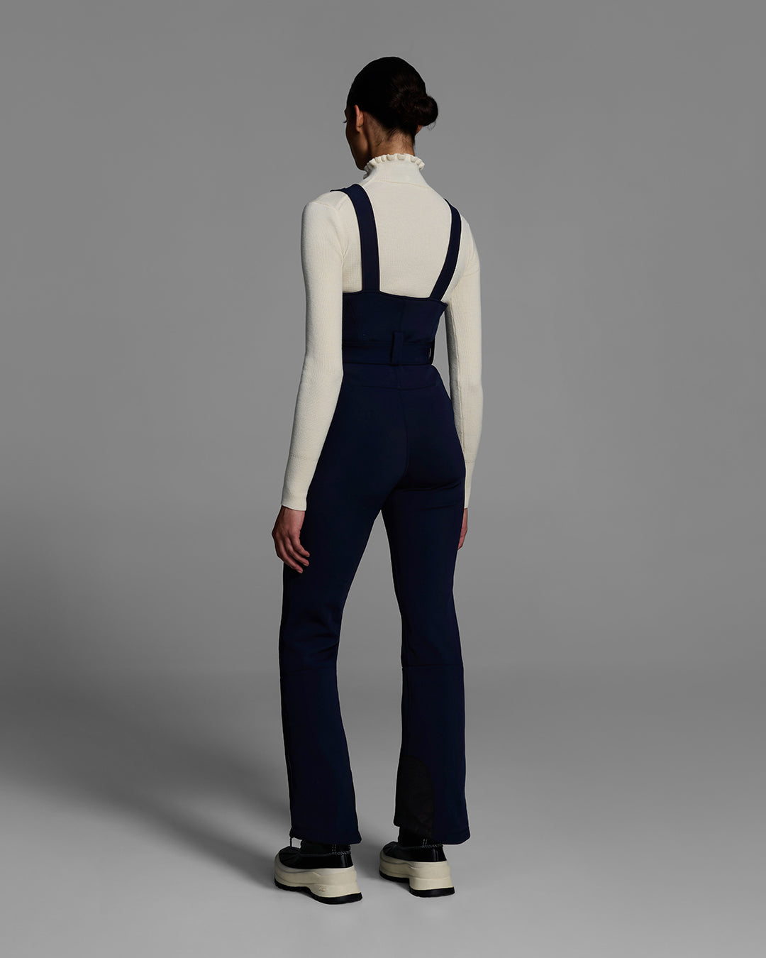 Back view of a female model wearing a belted indigo blue color ski bib with complementary white sweater. #color_indigo