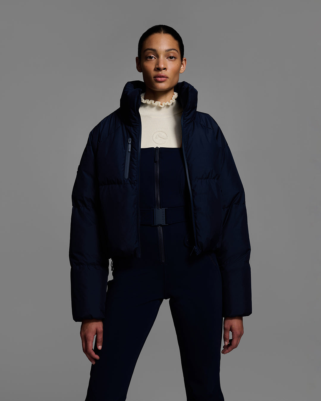 Female model wearing a belted indigo blue color ski bib with complementary white sweater and matching puffer ski jacket. #color_indigo