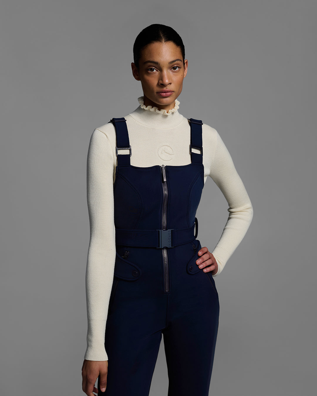 Detail shot of a female model wearing a belted indigo blue color ski bib with complementary white sweater. #color_indigo