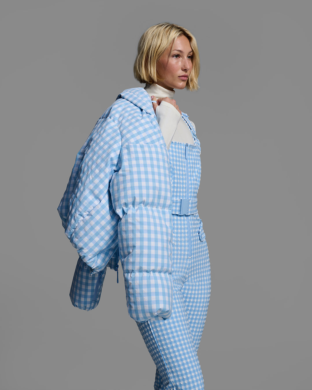 Female model wearing a belted cerulean blue color ski bib in a gingham print with a matching jacket hung over her shoulder. #color_cerulean gingham